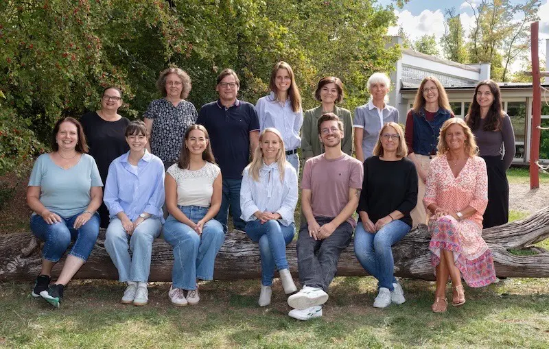 Gruppenfoto des Kollegiums auf einem Baumstamm vor dem Schulgebäude